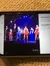 The image shows a group of performers on a stage, with some wearing red dresses or vests and black pants, underneath a spotlight and against a backdrop of blue stage lights, as viewed on a smartphone screen that also displays metadata about the photo.