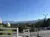 The image shows a clear blue sky over layered mountain ranges with lush greenery, viewed from a vantage point accented by white fences and flanked by pampas grass and a light pole, with hints of a parking area on the right.