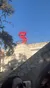 In the image, an individual wearing sunglasses is partially visible in the foreground, looking towards a bright red spiral sculpture situated on a building's terrace under a clear blue sky.