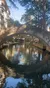 An arched stone bridge spans a reflective waterway lined with trees and buildings under a clear sky.