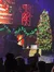 A musician is performing on stage beside a decorated Christmas tree, in a festive setting with an audience in the foreground.