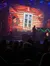 An audience watches a live performance with a festive stage backdrop showing a cozy log cabin window adorned with Christmas decorations.