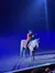 A performer dressed in an ornate costume with a cowboy hat is riding a horse in an arena under a spotlight, with an audience in the background.
