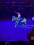 A person in a vibrant circus-like costume is riding a horse adorned with colorful stripes, performing for an audience in an arena with blue lighting.
