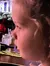 A young child looks on with interest at a vibrant theatrical performance while seated at a dining area with a plate of food in front of her.