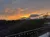 The image captures a vibrant sunset with orange-tinted clouds over a silhouette of rolling hills and trees, seen from a balcony adorned with string lights.