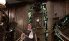 A person is smiling while standing on an ornate staircase adorned with festive Christmas decorations, flanked by elaborate wooden panels and a statuette.