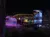 The image shows a vibrant night scene with a structure adorned with colorful lights and reflections on the water, creating a festive atmosphere.