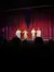 Performers take a bow on stage in front of a red curtain and an audience, suggesting the end of a theatrical show or performance.