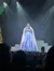 A performer in an elegant blue gown sings on stage amid a setting with a festive atmosphere, illuminated by beams of light which mimic a starry backdrop.
