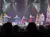 Five performers in sparkly dresses are singing and dancing on a festively decorated stage with Christmas trees and colorful lights.