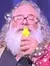 A performer with a long white beard is holding a microphone and standing next to a colorful puppet on a stage with blue curtains and festive decorations along the edge.