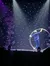 A performer is spinning inside a large hoop on stage under a spotlight, with a whimsical North Pole sign and a backdrop of lights suggesting a festive or winter-themed show.