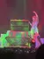 A performer in a sparkling outfit is gesturing dramatically on stage next to a large, brightly-lit chest and shimmering decorations.