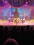 Performers dressed in colorful costumes are dancing on stage in a holiday-themed show with a festive backdrop and an audience watching.