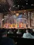 The image captures a vibrant theatrical performance with actors on stage and faux snow falling, as seen from the audience's perspective.