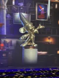 This image features a metallic statue of an animated character with a pointed hat, gesturing towards the viewer, placed on a pedestal within a room filled with shelves of various objects, under ambient lighting with a portrait of a character in a red cloak in the background.