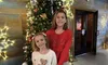 Two smiling children are standing in front of a decorated Christmas tree with presents at the base.