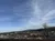 The image showcases a scenic overlook of a developed area with buildings, parking lots and roads, nestled among rolling hills under a vast blue sky adorned with wispy clouds.
