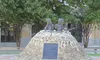 The image shows a memorial with sculptures and an informational plaque dedicated to the Korean War, set amidst a peaceful outdoor setting with trees in the background.