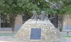 The image shows a memorial with sculptures and an informational plaque dedicated to the Korean War, set amidst a peaceful outdoor setting with trees in the background.