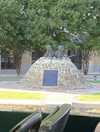 The image shows a memorial with sculptures and an informational plaque dedicated to the Korean War, set amidst a peaceful outdoor setting with trees in the background.