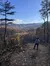 A person stands on a forest trail admiring a scenic view of mountains and autumn foliage under a clear blue sky.
