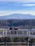 Two people stand on an elevated wooden deck overlooking a scenic view of layered mountains under a blue sky.