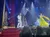 The image shows a theatrical performance with dancers in colorful costumes on a decorated stage, featuring Christmas trees and festive lighting.