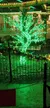 A tree covered in vibrant green lights stands behind a black fence, surrounded by festive decorations with a backdrop of a brightly lit area.