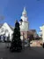 A public square features a decorated Christmas tree, surrounded by quaint buildings and a prominent clock tower under a clear blue sky.