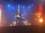 A performer is on stage with lighting effects, a background of machinery, and a burst of flames.