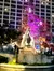 A stone monument stands in front of a tall building, with a tree covered in colorful Christmas lights nearby.