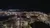 The image shows a nighttime view from a ski lift or aerial tramway overlooking a brightly lit town with various buildings and streets below.