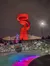 A large red, spiraling sculpture is illuminated at night above a stone bridge decorated with holiday lights and a wreath, with people gathered nearby.