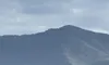 The image shows a mountain range under a cloudy sky with a few green trees in the foreground.
