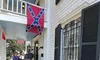 The image shows a porch with a Confederate flag hanging, and two people and a dog near the entrance of a building with white siding and black shutters.