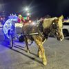 A horse pulls a carriage with bright lights, driven by someone dressed as the Grinch at night.