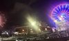 The image shows a colorful Ferris wheel and vibrant fireworks lighting up the night sky near a building and parked vehicles.