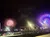 The image shows a colorful Ferris wheel and vibrant fireworks lighting up the night sky near a building and parked vehicles.