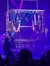 Two performers are hanging upside down in a metal structure on stage, with colorful lights and an audience watching.