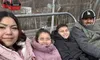 A family dressed warmly in coats is seated on a ski lift, with a red gondola visible in the background.
