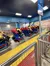 People are seated on a stationary amusement park ride with brightly colored seats and a sign indicating larger seats are available.