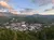 A small town nestled in a lush green valley is surrounded by forested mountains under a sky with scattered clouds.