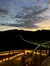The image shows a scenic view of a long, illuminated bridge stretching over a dark landscape at sunset, with a cloudy sky and hills in the background.