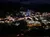 The image shows a nighttime view of a brightly lit cityscape seen from a high vantage point, possibly from a cable car or ski lift, with buildings and streets illuminated against a dark mountainous backdrop.