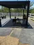 A group of alpacas is resting under a shaded structure surrounded by a white fence on a sunny day.