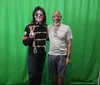 Two people are standing in front of a green screen, with one person dressed in a military-style black jacket making a peace sign.