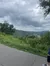 The image shows a cloudy sky over a lush, green landscape with a distant view of a town or city nestled among the hills, visible from a paved pathway.