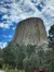 A massive, columnar rock formation rises dramatically from a forested area under a partly cloudy sky.